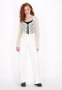 Häkelstrick-Cardigan in Off-White mit langen Ärmeln, einem runden Ausschnitt und Frontbindern, kombiniert mit einem schwarzen Oberteil und weißen Hosen.