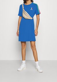 Modré bavlněné šaty ve formě trička s krátkým rukávem a bočními rozparky, s multibarevnými pruhy a logem Adidas. Béžová crossbody taška a bílé tenisky.