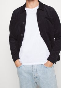 Svart cordjacka med knappstängning, två framfickor och krage, bärs över en vit t-shirt och ljusa denimjeans.