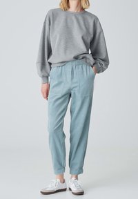Personne portant un sweat-shirt gris ample, un pantalon en velours côtelé bleu clair avec des poches, et des baskets blanches, se tenant devant un fond clair.