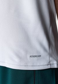 Witte sportieve mouwloze shirt met een geperforeerde textuur, met een Aeroready-logo aan de onderkant. Het shirt is gecombineerd met groene shorts.