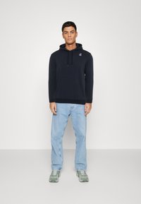 Felpa scura di colore navy in tessuto morbido con tasca frontale e cappuccio con cordino; abbinata a jeans azzurri e scarpe da ginnastica verdi.