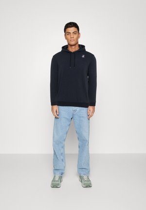 Tume sinine kapuutsiga sviiter pehmest kangast, millel on ees tasku ja nööriga kapuuts; kokku sobitatud helesiniste teksade ja roheliste tossudega.