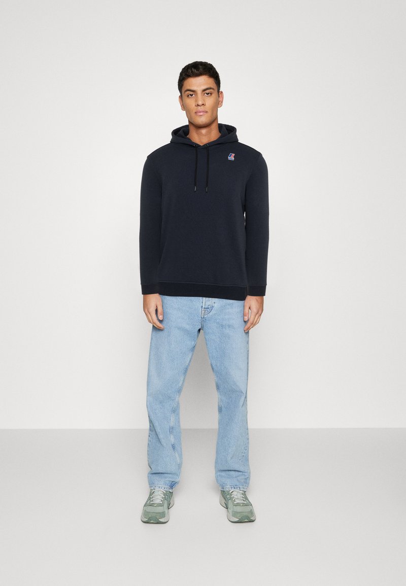 Felpa scura di colore navy in tessuto morbido con tasca frontale e cappuccio con cordino; abbinata a jeans azzurri e scarpe da ginnastica verdi.