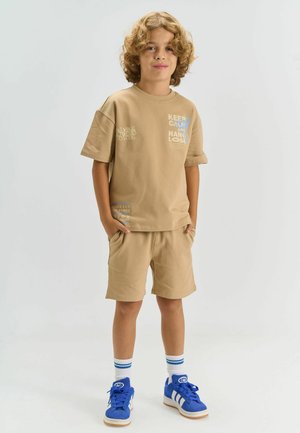 Ung dreng med krøllet hår står med hænderne i lommerne iført beige matchende shorts og t-shirt med blå sneakers og hvide sokker.