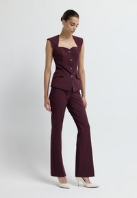 Completo su misura borgogna con scollo a carré, cinque bottoni e tasche frontali. Abbinato a scarpe con tacco alto bianche. Design elegante e strutturato.