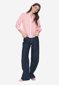 Roze blouses met oogjes en pofmouwen, knoopsluiting aan de voorkant. Gecombineerd met donker denim wijd uitlopende jeans en lichtroze schoeisel.