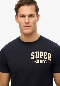 Camiseta de algodón navy con cuello redondo, que presenta un logo bordado en crema de "SUPER" y el texto "JPN DRY ATH" debajo, de mangas cortas.