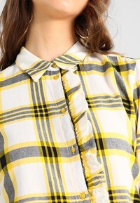 Camisa de cuadros amarillos y negros con cuello de solapa, tapa de botones con volantes y botones amarillos. Hecha de una tela ligera.