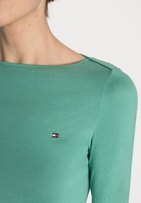 Mintgrünes Langarmshirt mit rundem Halsausschnitt, ausgestattet mit einem kleinen, mehrfarbigen Logo auf der Brust und einer glatten, dehnbaren Textur.