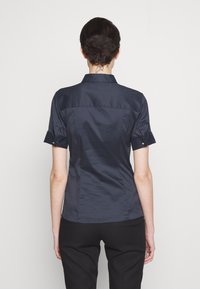 Marinblå kortärmad skjorta med krage, slät textur, figursydd design och uppvikta ärmar. Har en sömlös baksida och minimala sömnadsdetaljer.