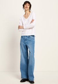 Frau mit dunklen Haaren, die ein weißes Langarmoberteil, hellblaue weitgeschnittene Jeans und schwarze Schuhe trägt, steht mit verschränkten Armen.