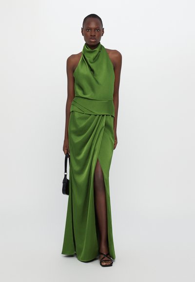 Green satin gown with a high neck, layered bodice, and a side slit. Model holds a small black handbag, wearing black strappy sandals.