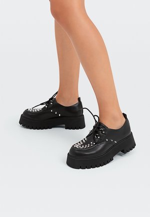 Chaussures en cuir noir avec une semelle épaisse, ornées de clous argentés sur les orteils et de surpiqûres décoratives. Conception à lacets avec bout rond.