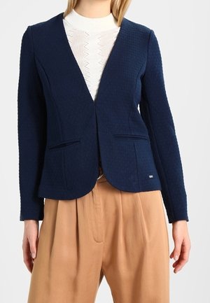 Femme portant un blazer bleu marine texturé sur un pull blanc à motifs et un pantalon ample taille haute couleur sable, sur un fond uni.