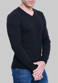 Pull léger noir avec col en V, manches longues et coupe ajustée. Le tissu semble lisse avec des poignets et un ourlet côtelés. Associé à un jean bleu ajusté.