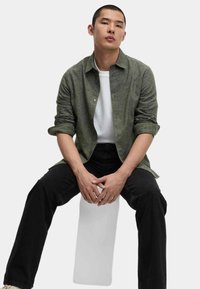 Camisa verde de botones hecha de tejido texturizado, con mangas remangadas, superpuesta sobre una camiseta blanca. Jeans negros de corte recto. Fondo neutro.