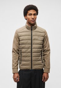 Veste matelassée taupe avec col montant, dotée d'une fermeture éclair sur le devant, de deux poches latérales et d'un patch logo sur la poitrine. Texture lisse.