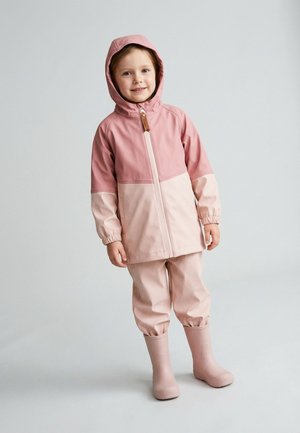 Young girl wearing a two-tone pink rain jacket with hood, matching pink rain pants, and pink rain boots, standing and smiling against a plain background.