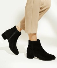 Black suede ankle boots with a block heel, featuring a side zip for closure and a smooth, soft texture. Paired with beige cropped pants.