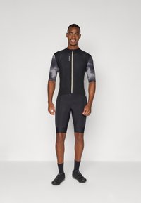 Sort cykeldragt med korte ærmer, der har et struktureret sort og gråt mønster, en lynlås foran, samt tætsiddende shorts. Sorte sko og sokker inkluderet.