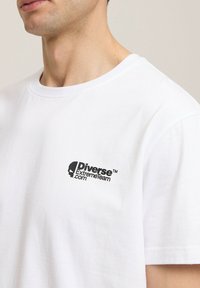 Biała koszulka bawełniana z okrągłym dekoltem, krótkimi rękawami i czarnym nadrukowanym logo na lewej piersi. Gładka faktura, casualowy design.