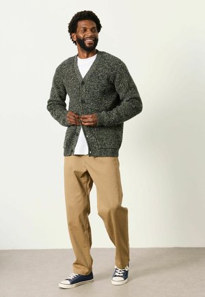 Mann lächelt, während er eine dunkelgraue Strickjacke über ein weißes Hemd knöpft, trägt beige Hose und marineblaue Sneaker, steht auf einem schlichten Boden.