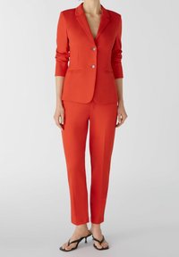 Costume sur mesure rouge comprenant une veste ajustée à deux boutons et un pantalon slim. Tissu lisse avec des manches longues et sans motifs.