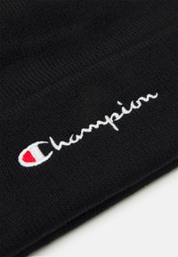 Gorro de punto negro con el logotipo "Champion" bordado en blanco y un acento rojo en la letra "C". Textura suave y diseño ajustado.
