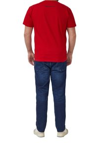 Mann steht mit dem Rücken zur Kamera, trägt ein rotes kurzärmeliges T-Shirt, blaue Jeans und weiße Turnschuhe vor einem weißen Hintergrund.