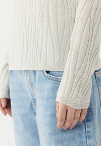 Pull en tricot couleur crème avec texture côtelée et manches longues, associé à un jean en denim bleu clair. Le pull présente un léger effet scintillant.
