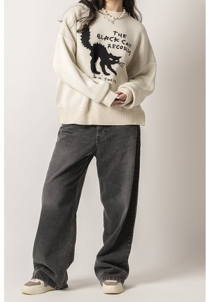 Femme portant un pull crème oversize avec un chat noir et un texte, assorti à un jean ample gris foncé et des baskets beiges.