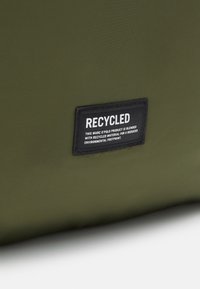 Zelený sáček z recyklovaného materiálu s obdélníkovou černou etiketou, na které je bílý text uvádějící jeho udržitelné složení a podrobnosti o značce.