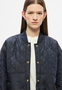 Tengerészkék steppelt bomber dzseki bordázott gallérral, arany patentgombokkal és texturált felülettel. Fehér felsővel viselve.
