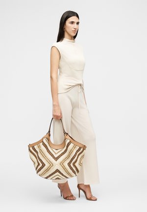 Frau in beige sleeveless Top und Hose hält große braun-weiße geflochtene Handtasche, trägt braune Sandalen mit hohen Absätzen, steht seitlich.