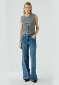 Grauer ärmelloser Knopf-Down-Weste kombiniert mit hellblauen, hoch taillierten, ausgestellten Jeans und dunkelburgunderfarbenen Pumps. Glatte Texturen.