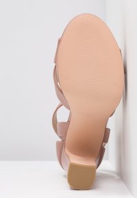 Semelle d'une sandale rose avec un talon bas et épais reposant contre une surface blanche, montrant une semelle texturée et des brides lisses.