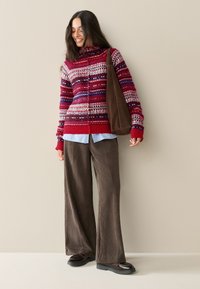 Kleurige gebreide trui met rood, blauw en witte patronen, gecombineerd met een lichtblauw overhemd en bruine wijde corduroy broek. Bruine tote tas.