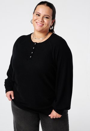 Glimlachende vrouw in een zwart lange mouwen top, met zilveren oorringen en meerdere kettingen, staand tegen een effen witte achtergrond.