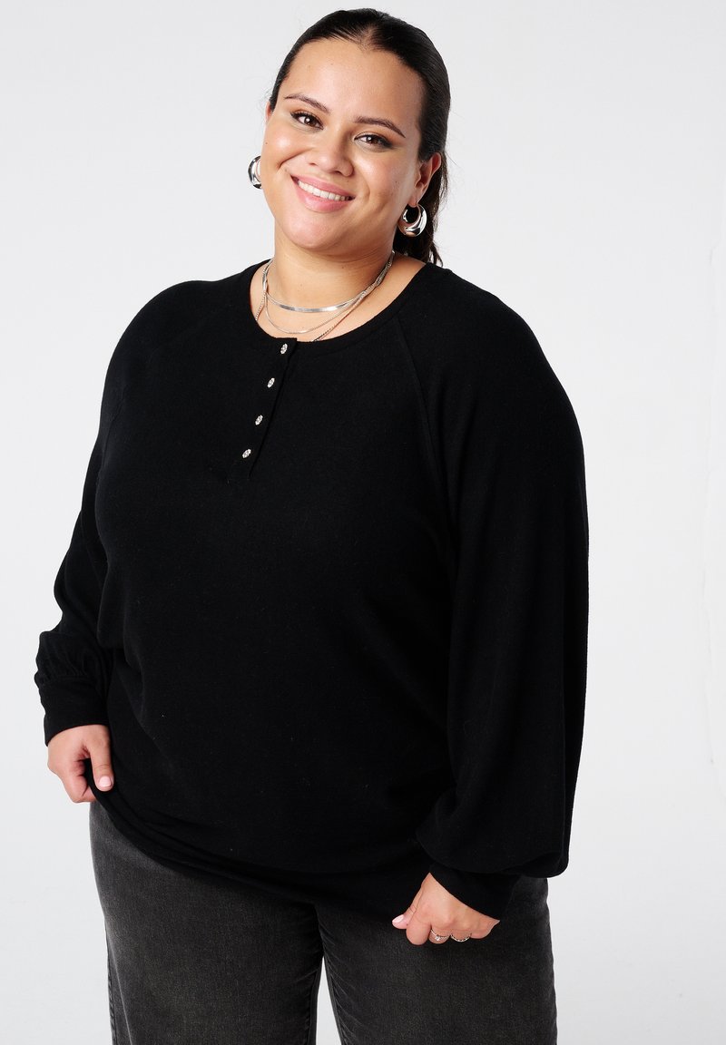 Glimlachende vrouw in een zwart lange mouwen top, met zilveren oorringen en meerdere kettingen, staand tegen een effen witte achtergrond.