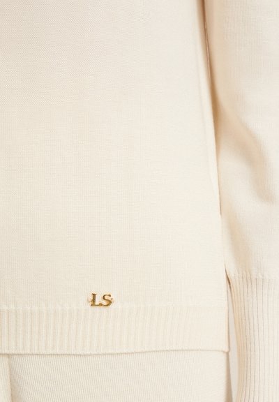 Beige neulepusero, jossa on nuohottu pinta ja kultainen "LS" -logo helmaa koristamassa. Suunnittelussa on sivuhalkiot lisäämään liikkumavaraa.