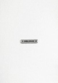 Fondo de tela blanca con una pequeña etiqueta de metal plateada que dice "SMILODOX" y presenta dos agujeros para sujeción. La etiqueta tiene un acabado suave.