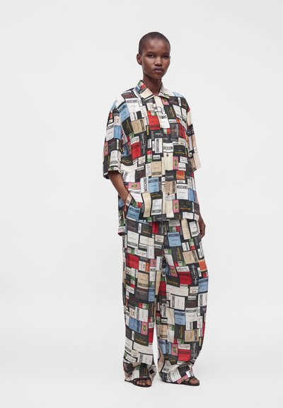 Le mannequin porte une chemise et un pantalon amples assortis imprimés avec des étiquettes colorées du logo Moschino sur fond blanc.