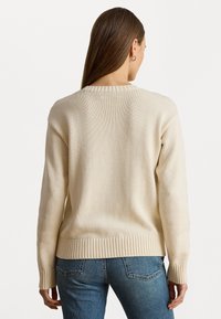 Pull en crème avec un tricot texturé, des poignets et un ourlet côtelés. Design classique à col rond, porté avec un jean bleu, montré de dos.
