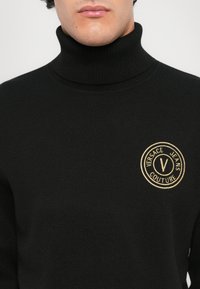 Moški v črnem puloverju s turtleneckom in z zlatim krožnim logotipom "Versace Jeans Couture" na levem prsnem delu.