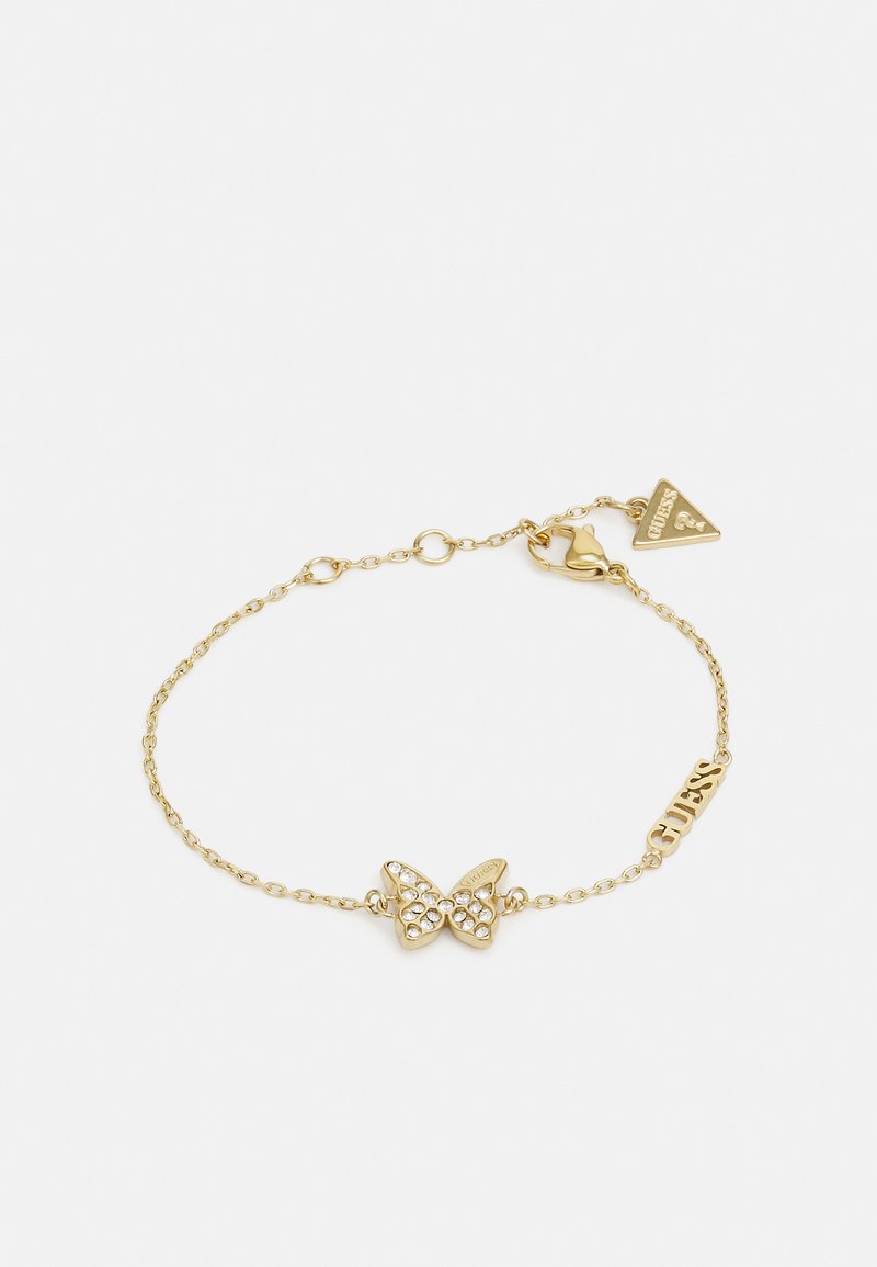 Pulsera de cadena de oro con un charm de mariposa adornado con piedras de strass y una pequeña etiqueta grabada con "GUESS". Diseño de eslabones simple.