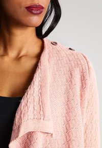 Helles rosa gestricktes Cardigan mit einem strukturierten Muster, Schalkragen und dekorativem Knopf an der Schulter. Weicher Stoff und lockere Passform.