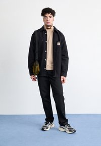 Jeune homme atteint de vitiligo portant une veste noire, une chemise beige, un pantalon noir, des baskets, et portant un sac à bandoulière vert olive, se tenant à l'intérieur.