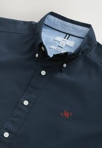 Granatowa koszula zapinana na guziki o slim fit, z białymi guzikami i małym, czerwonym haftowanym logo na piersi. Jasnoniebieskie paski wykończenia kołnierza.