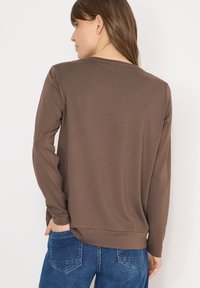 Chemise marron à manches longues, coupe décontractée et texture lisse, avec un col rond et un détail discret à l'ourlet. Associée à un jean bleu.
